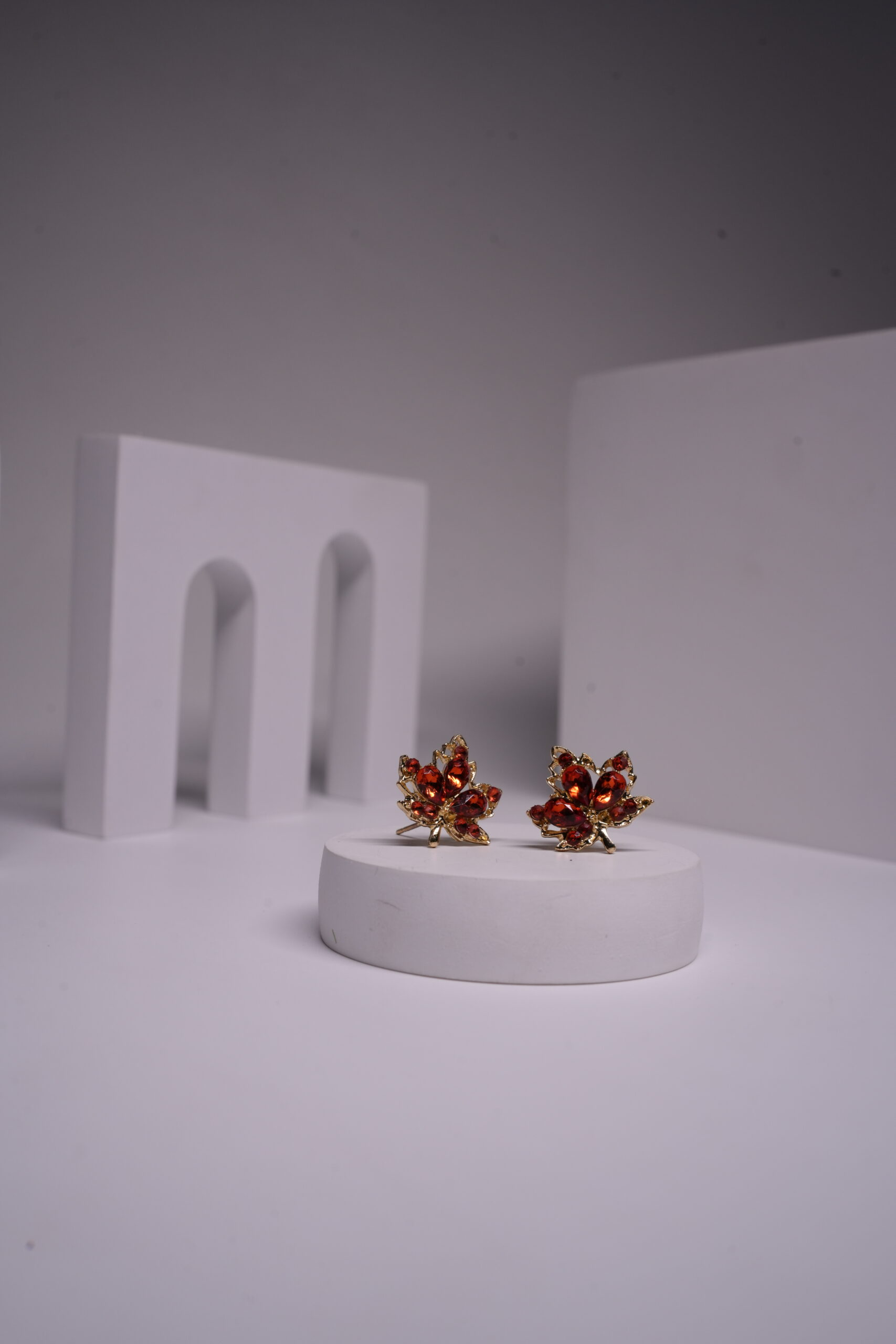 Red crystal maple leaf stud earrings with gold-toned metal frame, featuring deep red faceted stones in oval and round cuts, displayed on a white prop.