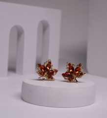 Red crystal maple leaf stud earrings with gold-toned metal frame, featuring deep red faceted stones in oval and round cuts, displayed on a white prop.