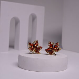Red crystal maple leaf stud earrings with gold-toned metal frame, featuring deep red faceted stones in oval and round cuts, displayed on a white prop.