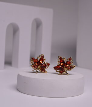 Red crystal maple leaf stud earrings with gold-toned metal frame, featuring deep red faceted stones in oval and round cuts, displayed on a white prop.