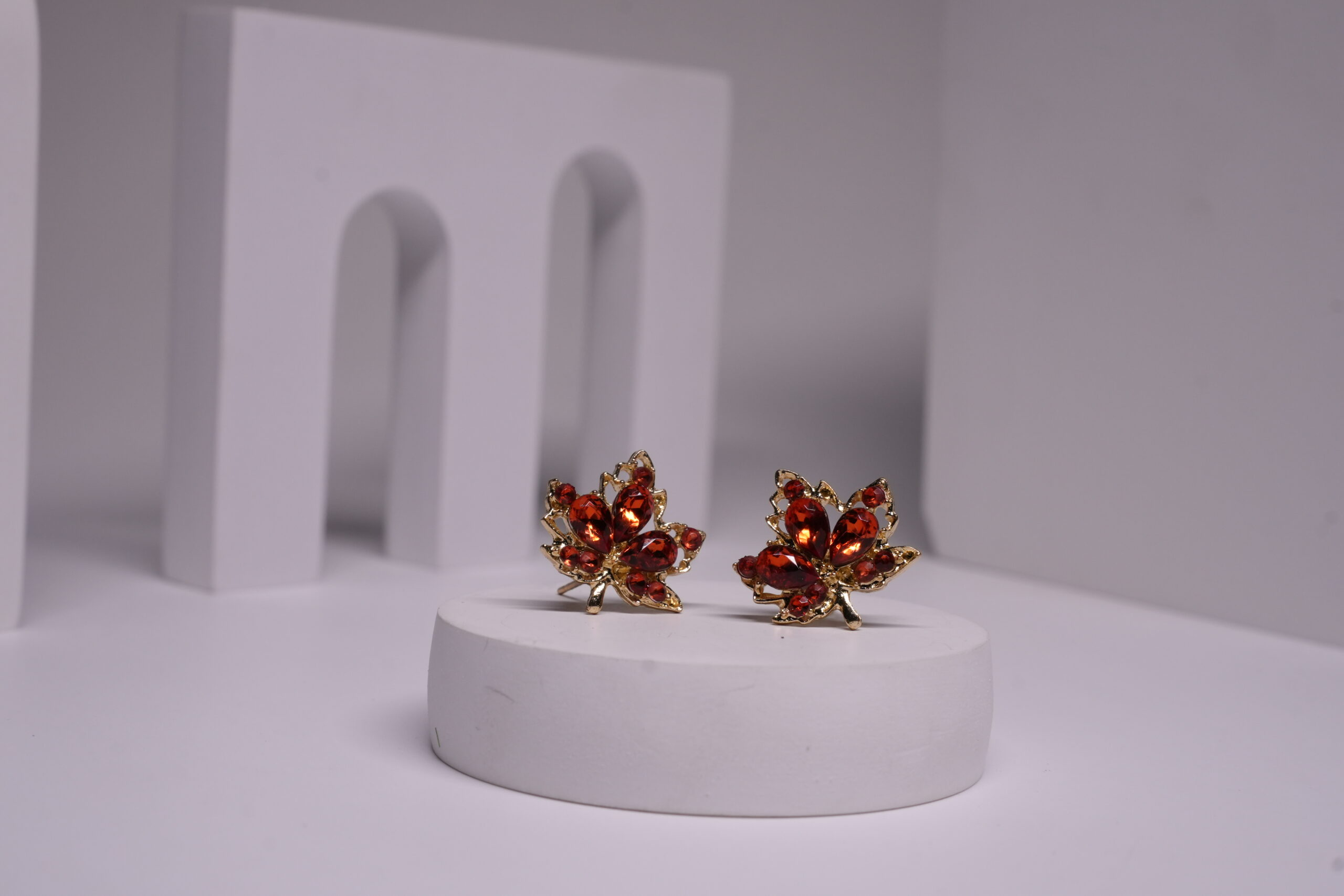Red crystal maple leaf stud earrings with gold-toned metal frame, featuring deep red faceted stones in oval and round cuts, displayed on a white prop.
