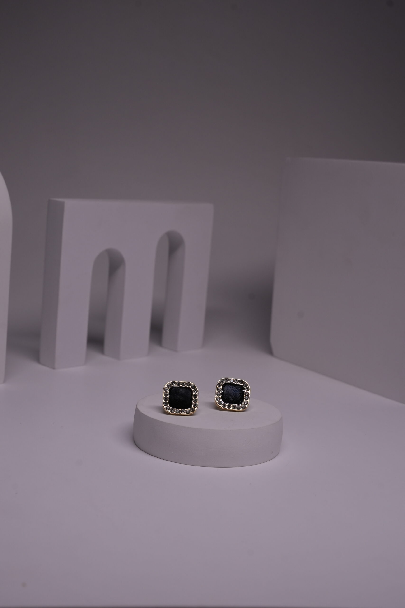 Black square stud earrings with pearl bead halo border in gold-toned frame, displayed as a pair on a white prop against a grey background.