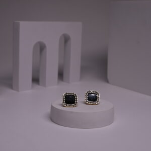 Black square stud earrings with pearl bead halo border in gold-toned frame, displayed as a pair on a white prop against a grey background.