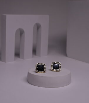 Black square stud earrings with pearl bead halo border in gold-toned frame, displayed as a pair on a white prop against a grey background.