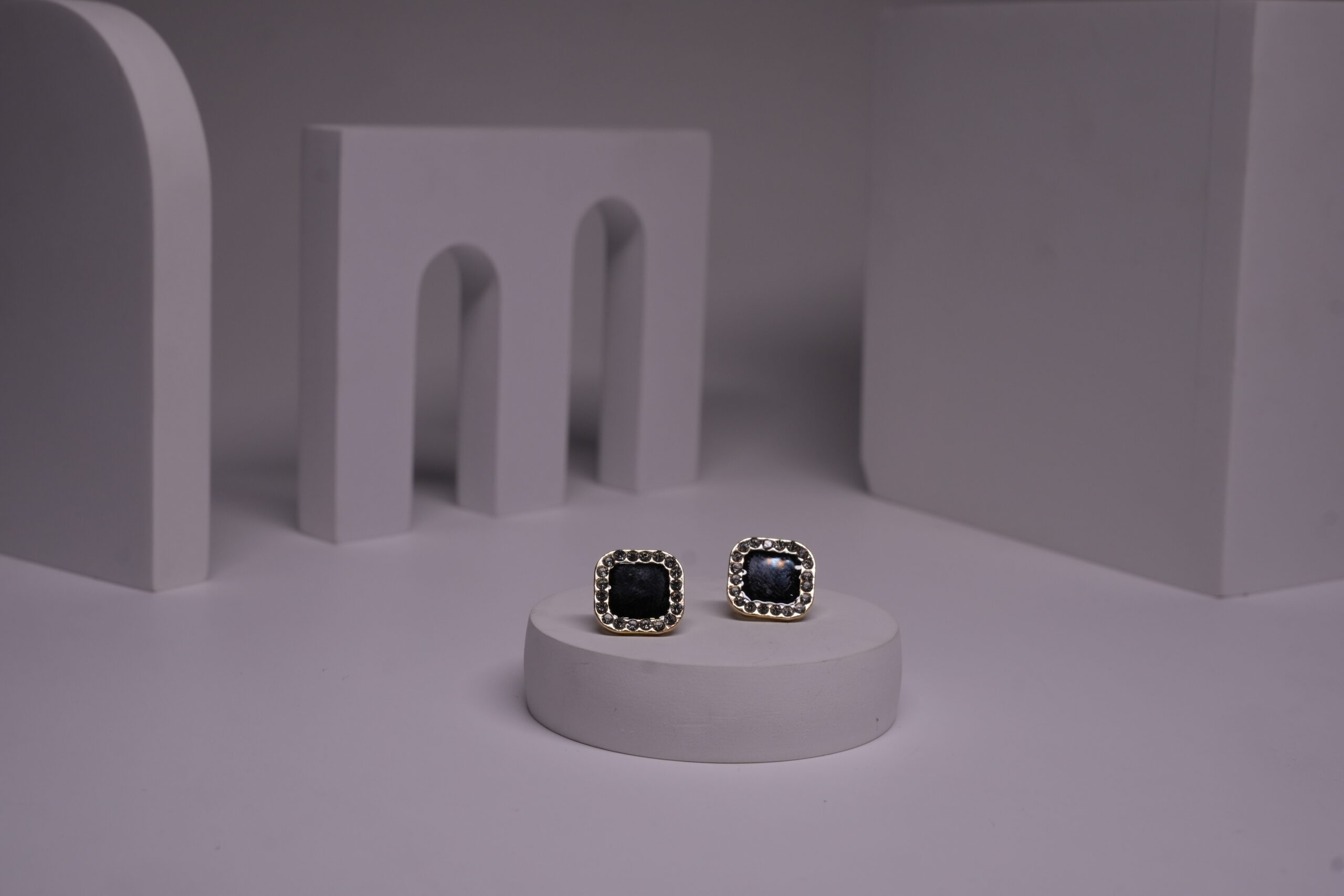 Black square stud earrings with pearl bead halo border in gold-toned frame, displayed as a pair on a white prop against a grey background.