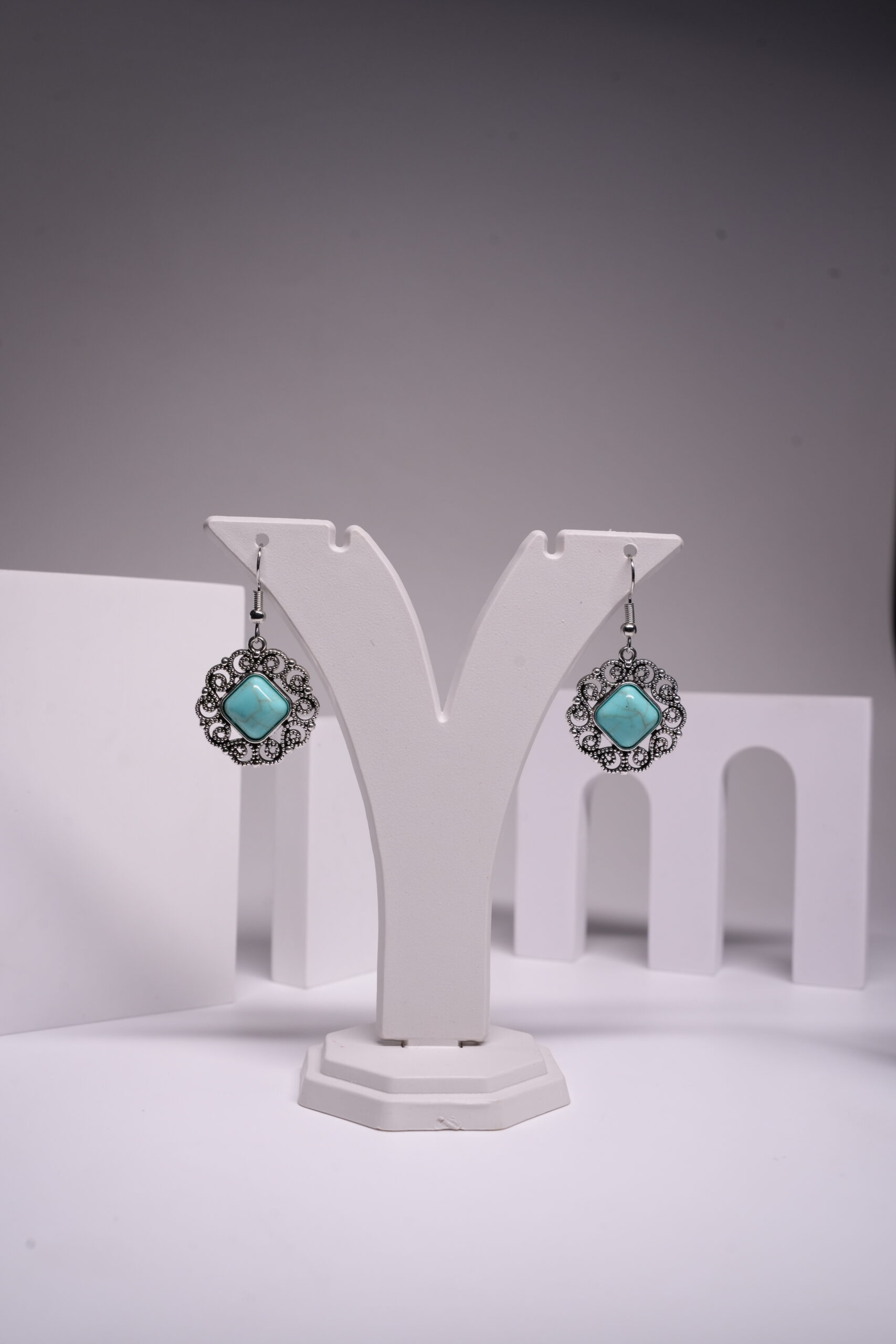 Oxidised silver-tone floral filigree drop earrings with square turquoise stone centre, displayed on a white earring stand