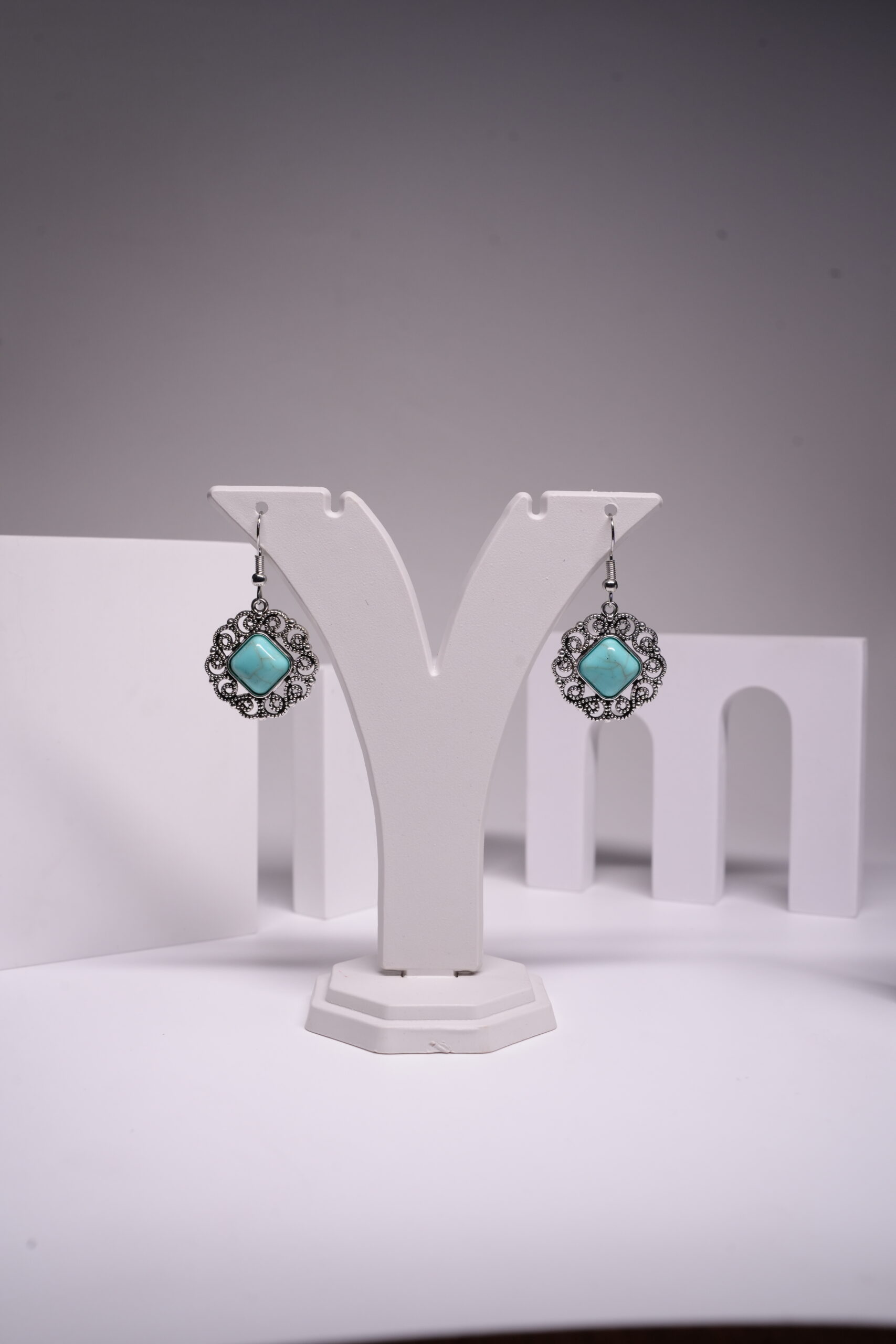 Oxidised silver-tone floral filigree drop earrings with square turquoise stone centre, displayed on a white earring stand