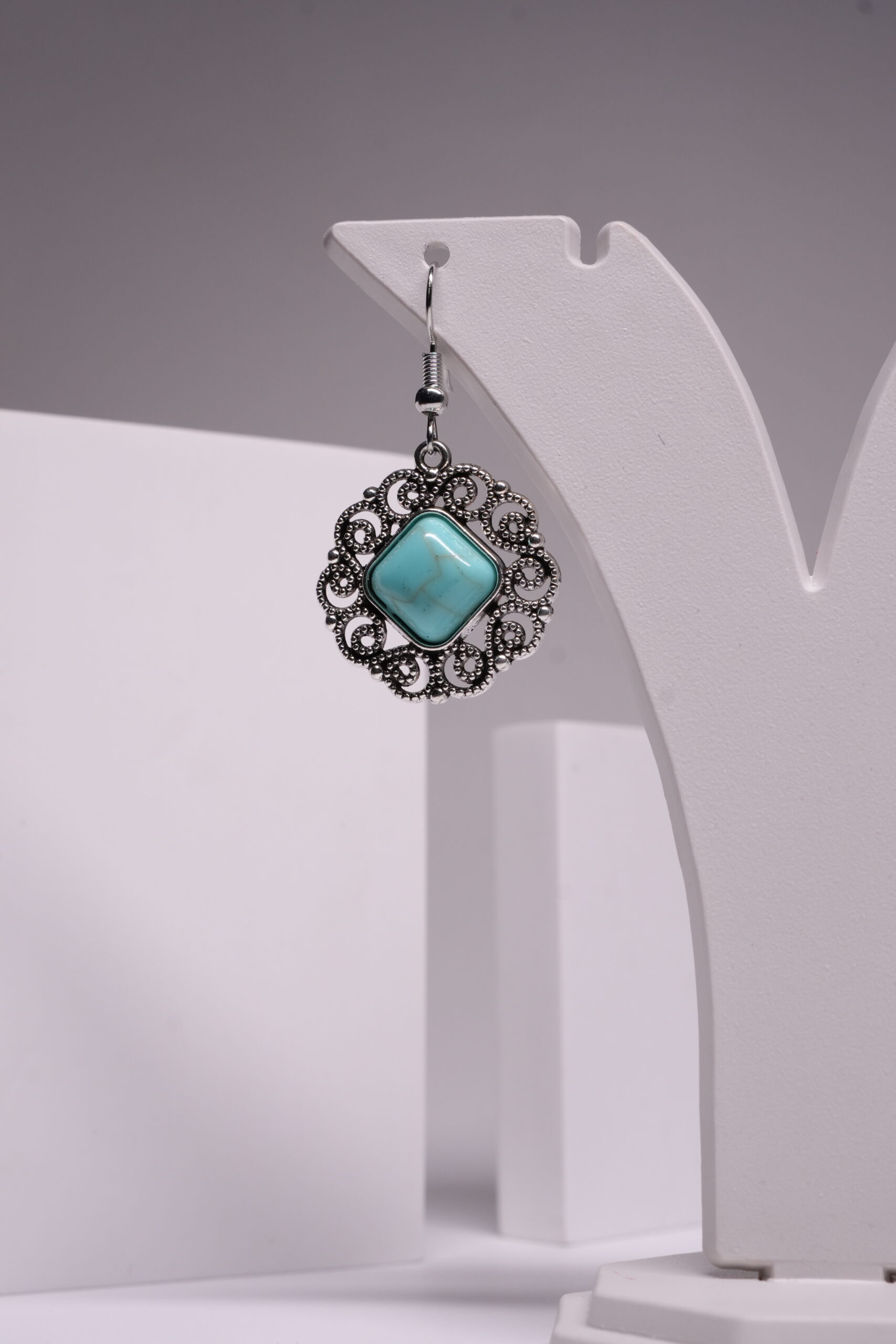 Oxidised silver-tone floral filigree drop earrings with square turquoise stone centre, displayed on a white earring stand