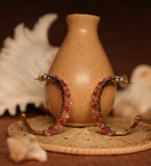 Pair of large gold-tone hoop earrings fully encrusted with pink and rose-red crystals in an ombre gradient, displayed upright against a ceramic vase with seashell styling props.