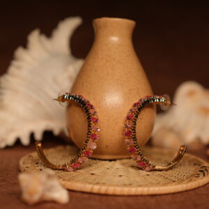 Pair of large gold-tone hoop earrings fully encrusted with pink and rose-red crystals in an ombre gradient, displayed upright against a ceramic vase with seashell styling props.