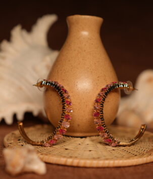 Pair of large gold-tone hoop earrings fully encrusted with pink and rose-red crystals in an ombre gradient, displayed upright against a ceramic vase with seashell styling props.