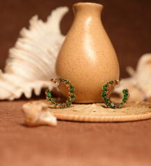 Pair of small gold-tone hoop earrings fully encrusted with vivid emerald green crystals in prong settings, displayed leaning against a speckled ceramic vase on a wicker coaster with seashells in the background.