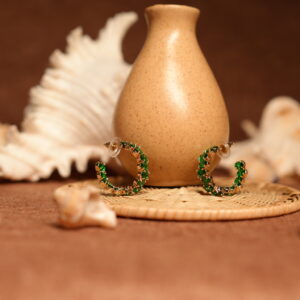 Pair of small gold-tone hoop earrings fully encrusted with vivid emerald green crystals in prong settings, displayed leaning against a speckled ceramic vase on a wicker coaster with seashells in the background.