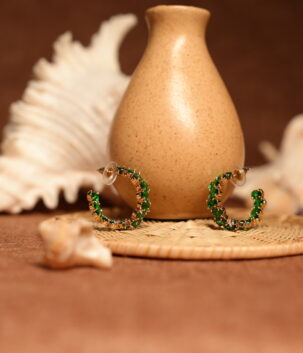 Pair of small gold-tone hoop earrings fully encrusted with vivid emerald green crystals in prong settings, displayed leaning against a speckled ceramic vase on a wicker coaster with seashells in the background.