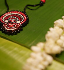 Handcrafted Theyyam face pendant necklace in red black and white with black beads on black thread cord displayed on banana leaf with jasmine flowers