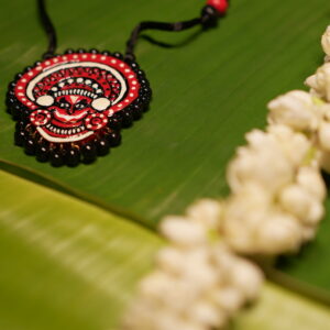 Handcrafted Theyyam face pendant necklace in red black and white with black beads on black thread cord displayed on banana leaf with jasmine flowers