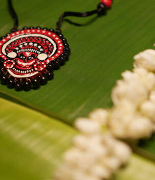 Handcrafted Theyyam face pendant necklace in red black and white with black beads on black thread cord displayed on banana leaf with jasmine flowers