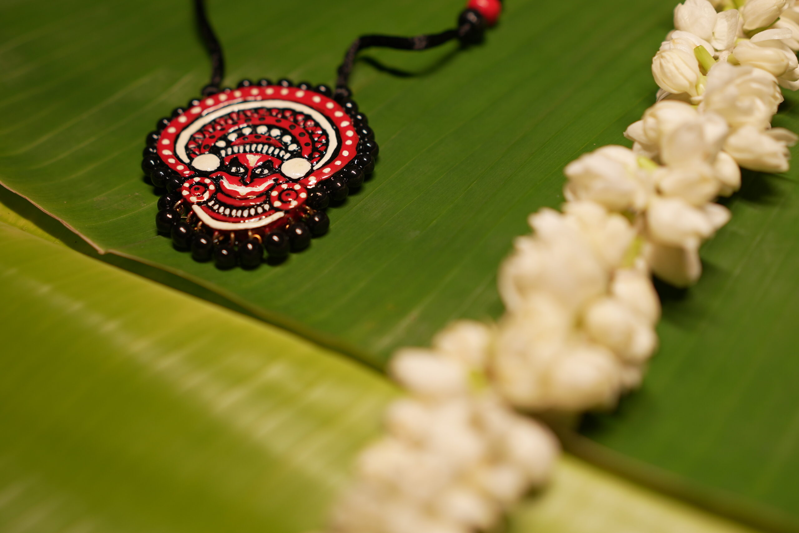 Handcrafted Theyyam face pendant necklace in red black and white with black beads on black thread cord displayed on banana leaf with jasmine flowers