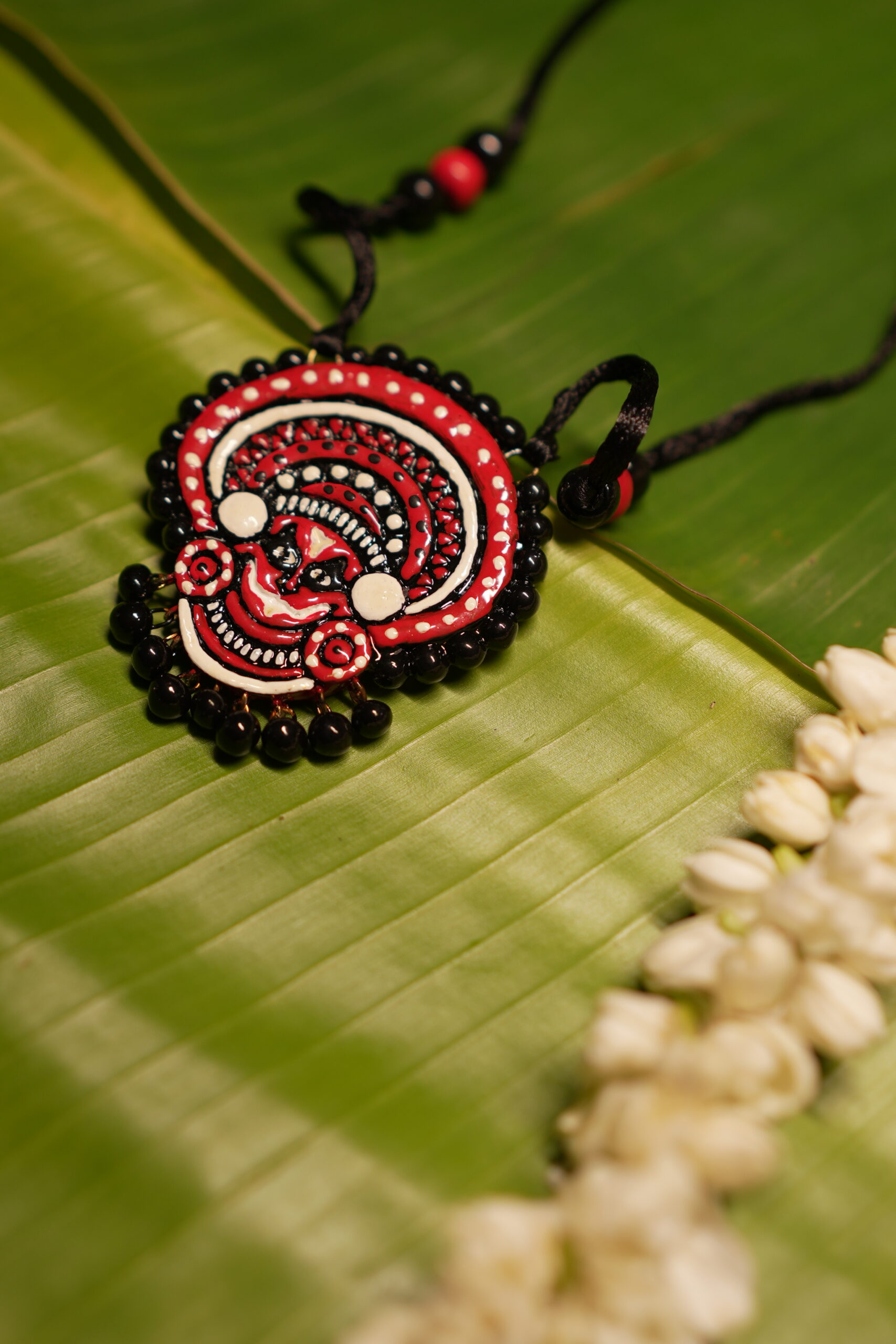 Handcrafted Theyyam face pendant necklace in red black and white with black beads on black thread cord displayed on banana leaf with jasmine flowers