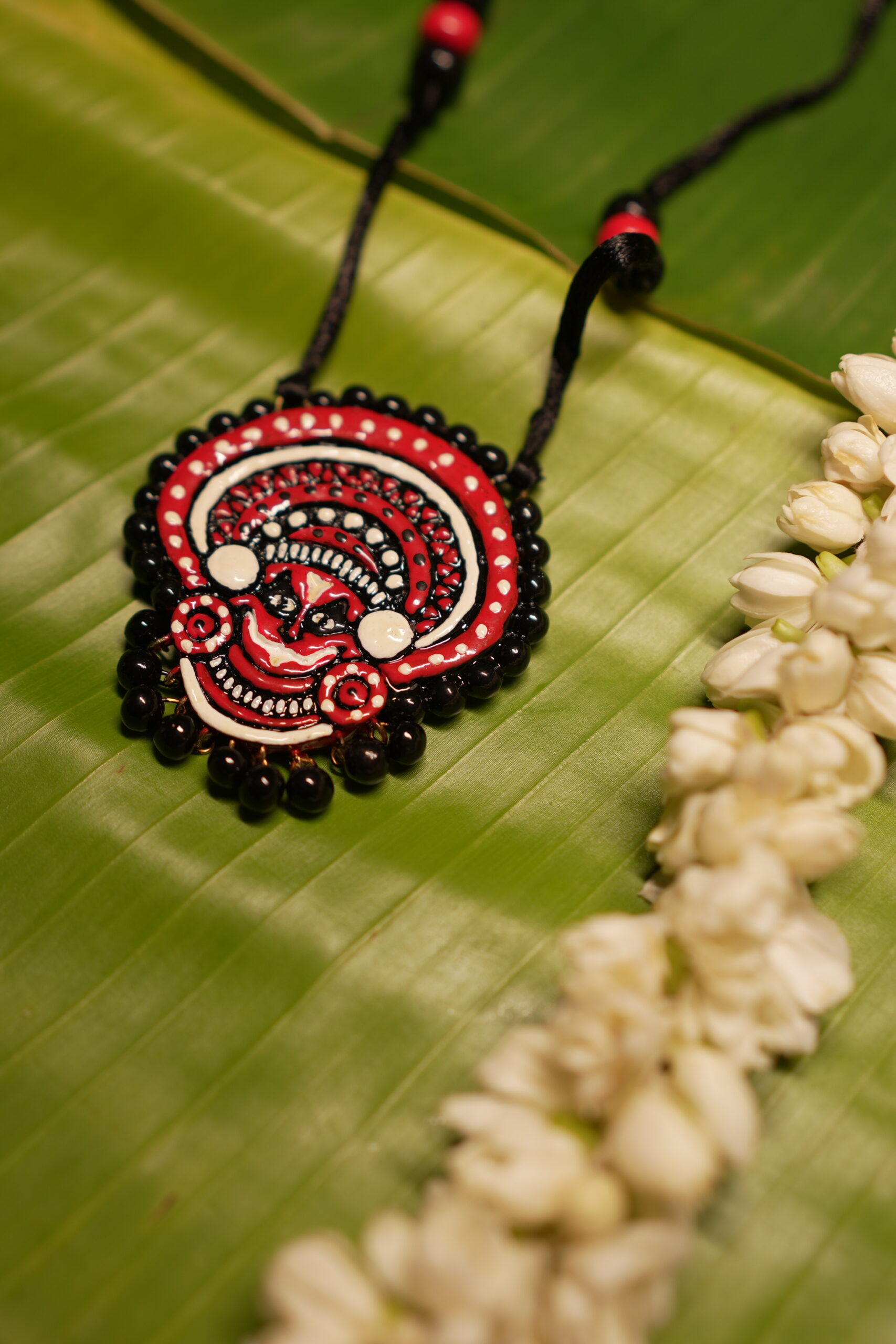 Handcrafted Theyyam face pendant necklace in red black and white with black beads on black thread cord displayed on banana leaf with jasmine flowers