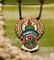 Handcrafted Kathakali face pendant necklace in green and gold with multicolor beads on black thread cord, displayed on banana leaf with jasmine flowers