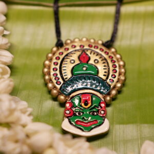 Handcrafted Kathakali face pendant necklace in green and gold with multicolor beads on black thread cord, displayed on banana leaf with jasmine flowers