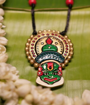 Handcrafted Kathakali face pendant necklace in green and gold with multicolor beads on black thread cord, displayed on banana leaf with jasmine flowers