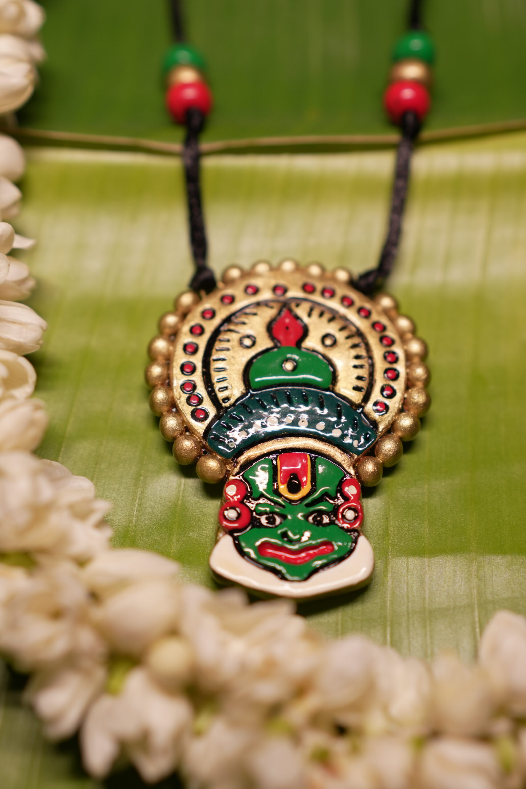 Handcrafted Kathakali face pendant necklace in green and gold with multicolor beads on black thread cord, displayed on banana leaf with jasmine flowers