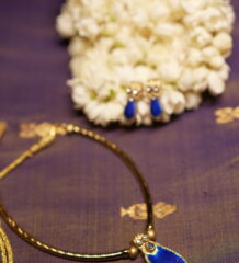 Gold-tone Kundan necklace with royal blue teardrop stone pendant and matching blue drop earrings displayed on purple silk fabric with white jasmine flowers