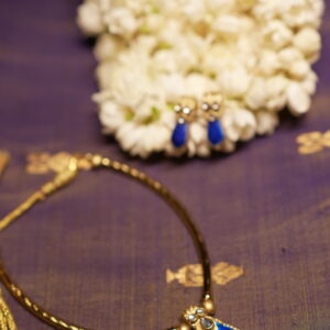 Gold-tone Kundan necklace with royal blue teardrop stone pendant and matching blue drop earrings displayed on purple silk fabric with white jasmine flowers
