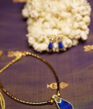 Gold-tone Kundan necklace with royal blue teardrop stone pendant and matching blue drop earrings displayed on purple silk fabric with white jasmine flowers