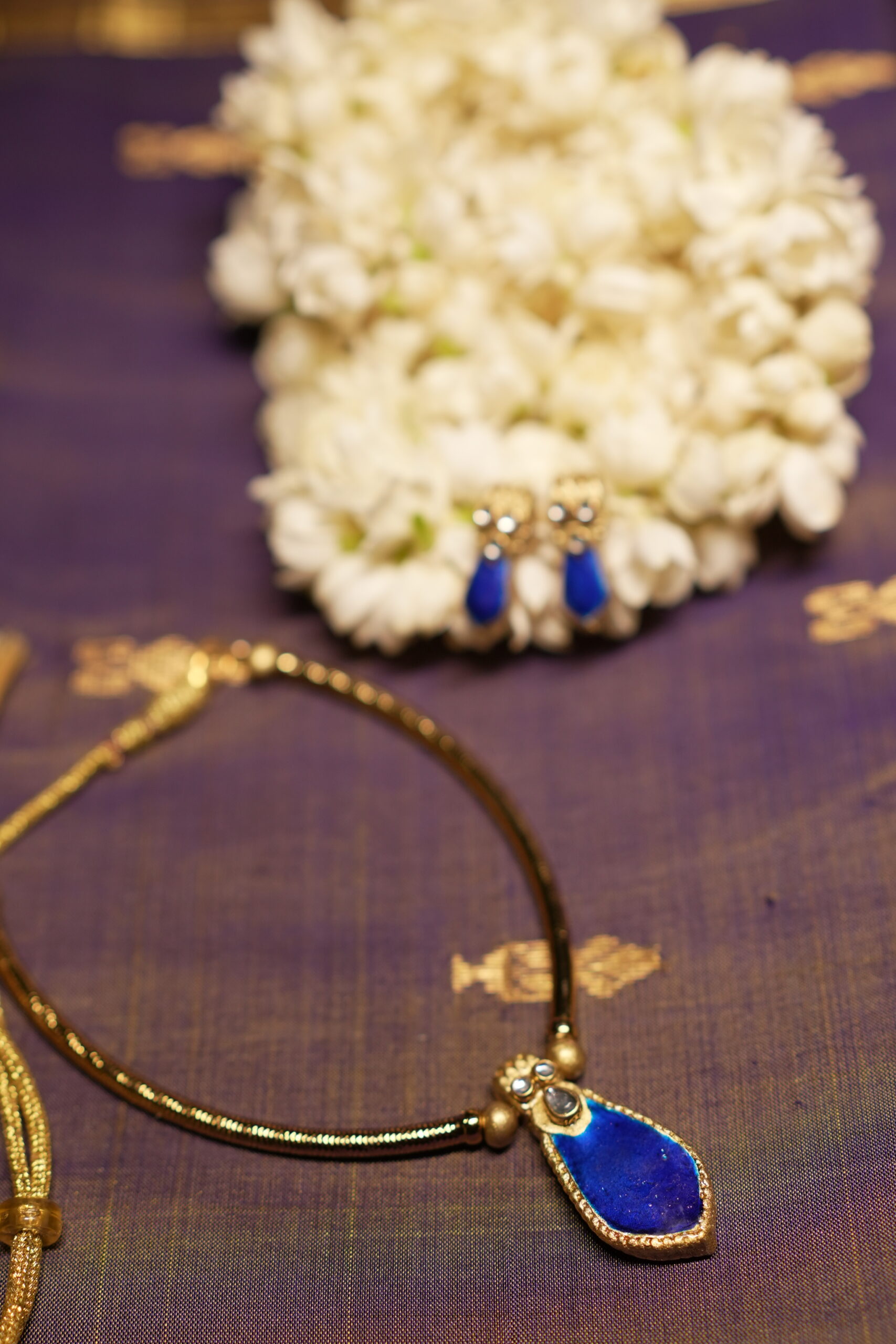 Gold-tone Kundan necklace with royal blue teardrop stone pendant and matching blue drop earrings displayed on purple silk fabric with white jasmine flowers