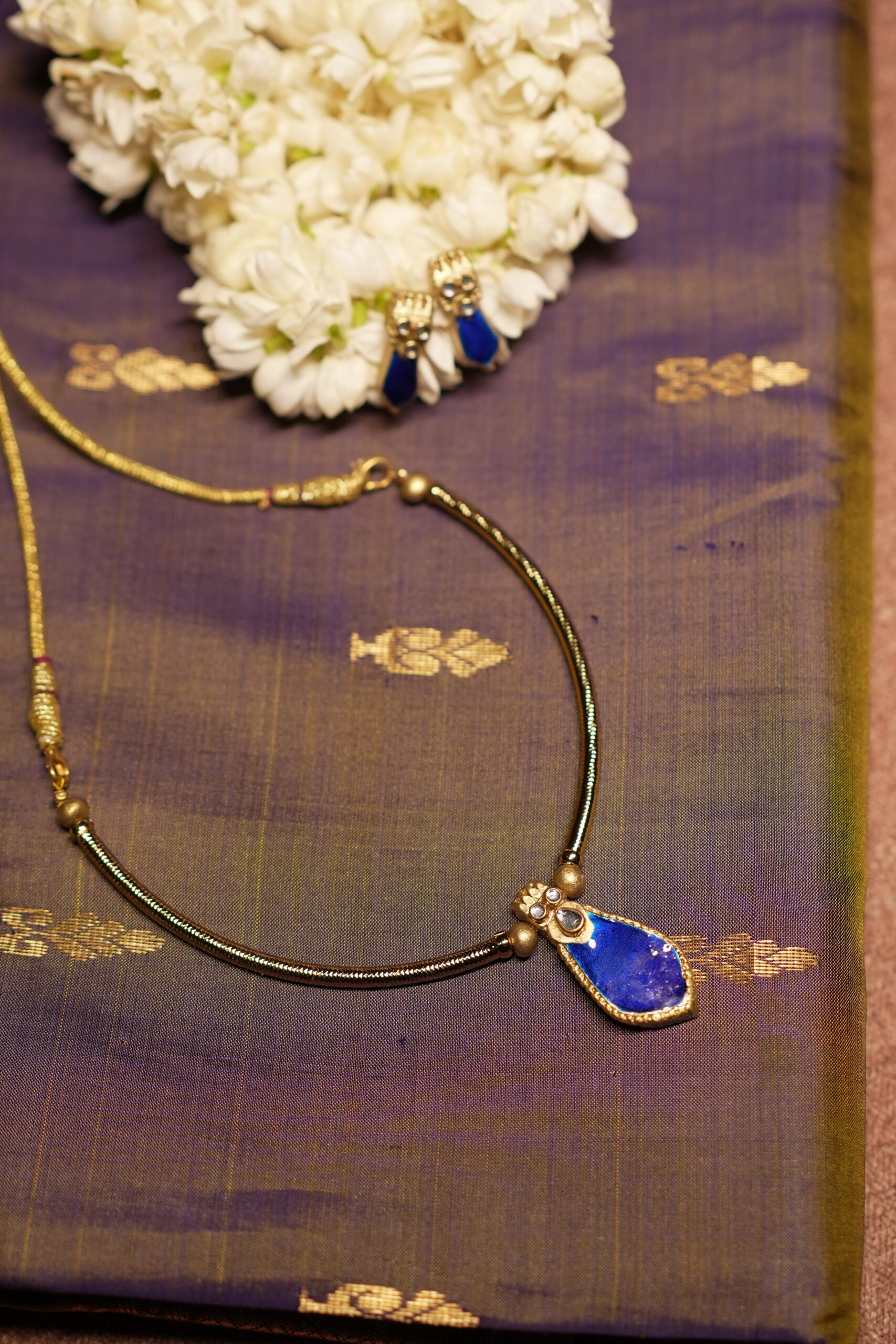 Gold-tone Kundan necklace with royal blue teardrop stone pendant and matching blue drop earrings displayed on purple silk fabric with white jasmine flowers