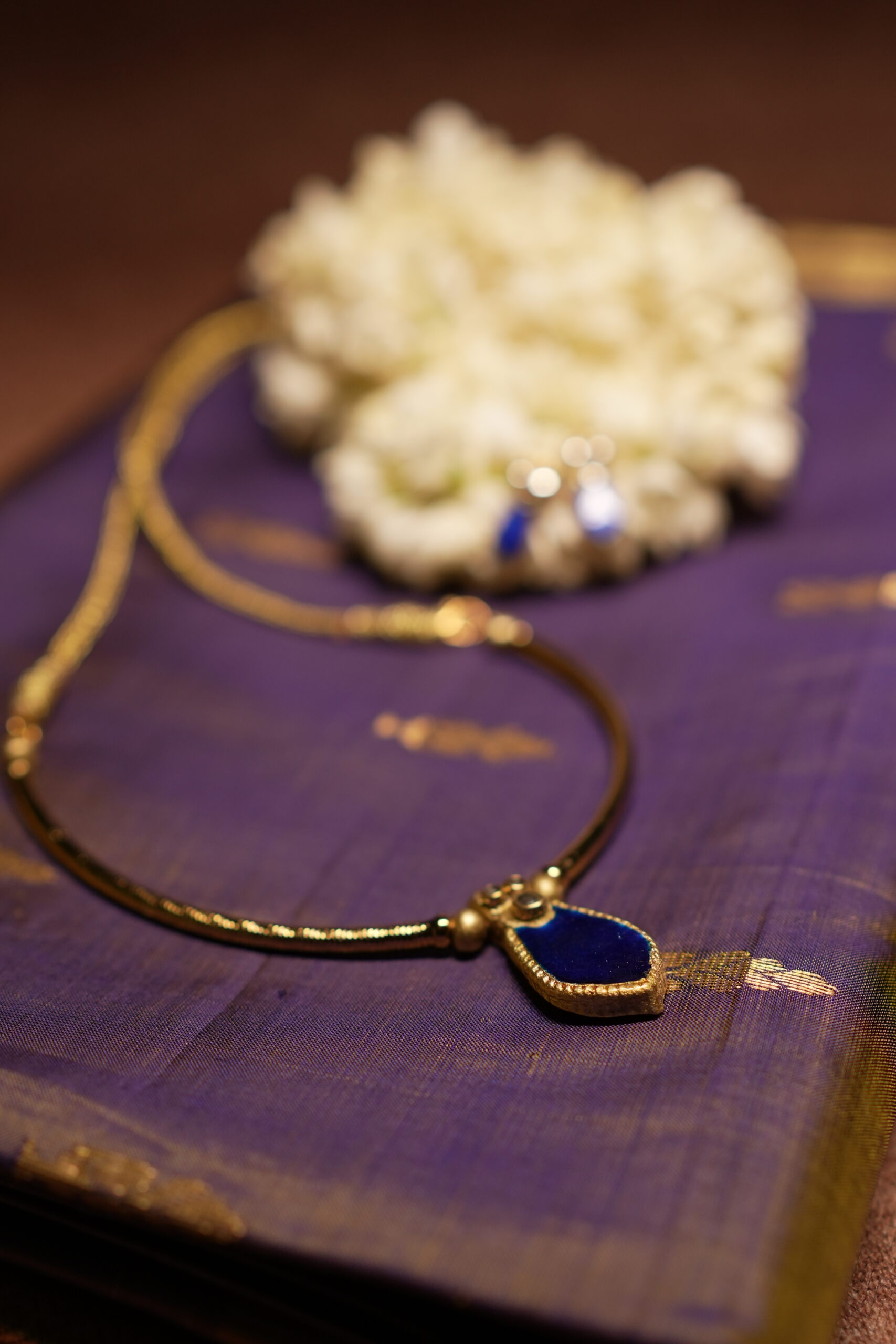 Gold-tone Kundan necklace with royal blue teardrop stone pendant and matching blue drop earrings displayed on purple silk fabric with white jasmine flowers