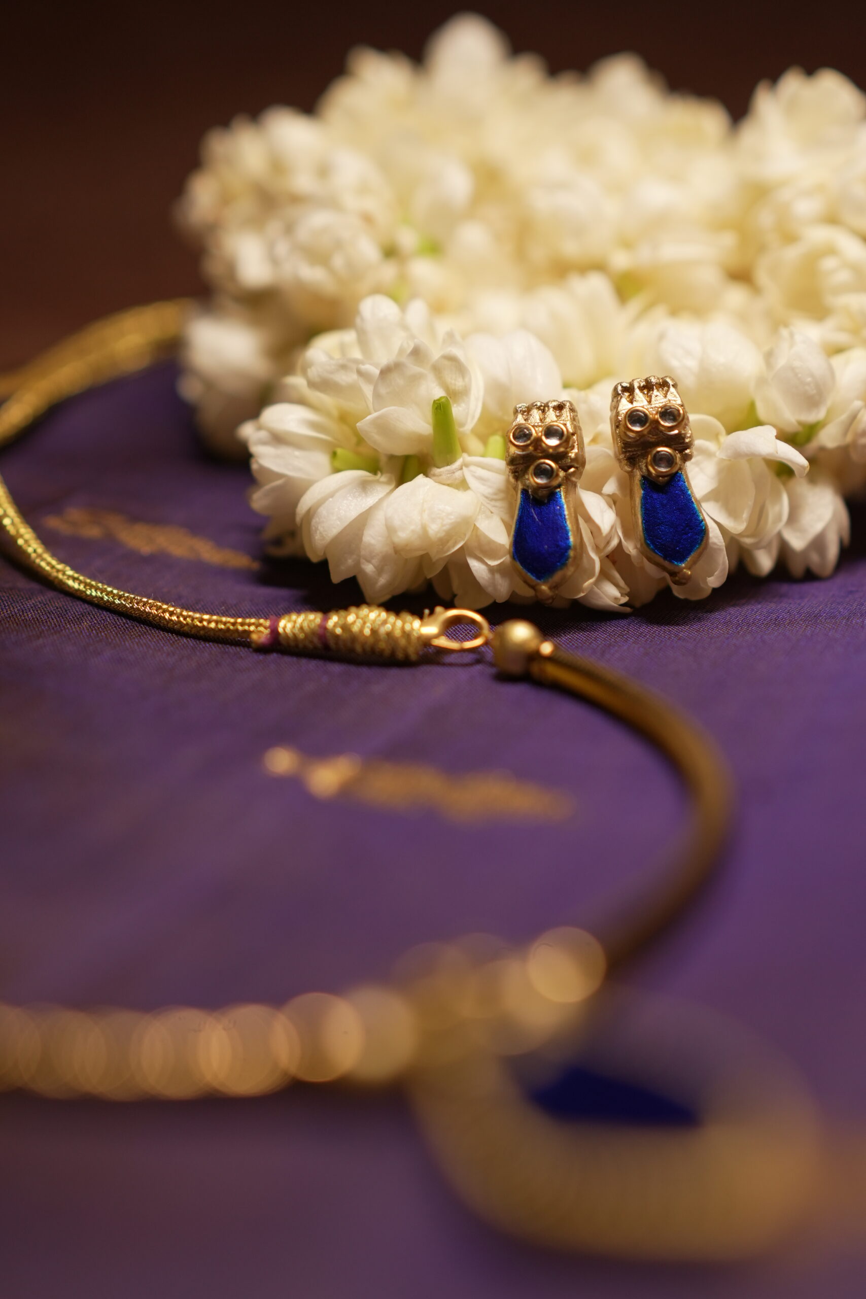 Gold-tone Kundan necklace with royal blue teardrop stone pendant and matching blue drop earrings displayed on purple silk fabric with white jasmine flowers