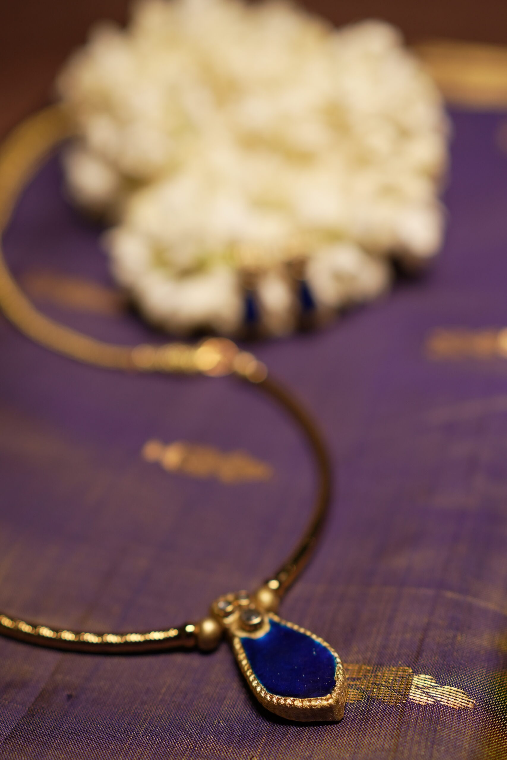 Gold-tone Kundan necklace with royal blue teardrop stone pendant and matching blue drop earrings displayed on purple silk fabric with white jasmine flowers
