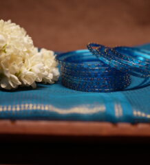 Translucent blue glass bangles with gold glitter detailing, set of six, displayed on a teal silk saree with white jasmine flowers in the background.