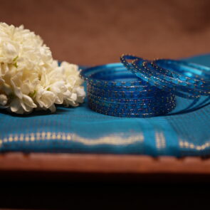 Translucent blue glass bangles with gold glitter detailing, set of six, displayed on a teal silk saree with white jasmine flowers in the background.
