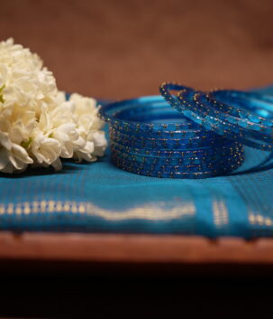 Translucent blue glass bangles with gold glitter detailing, set of six, displayed on a teal silk saree with white jasmine flowers in the background.