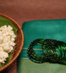 Deep emerald green glass bangles with rose gold glitter detailing, set of six, resting on a teal silk saree beside a wicker basket of fresh white jasmine flowers.