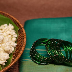 Deep emerald green glass bangles with rose gold glitter detailing, set of six, resting on a teal silk saree beside a wicker basket of fresh white jasmine flowers.