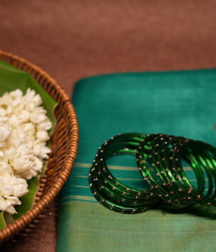 Deep emerald green glass bangles with rose gold glitter detailing, set of six, resting on a teal silk saree beside a wicker basket of fresh white jasmine flowers.