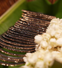 Set of six black glass bangles densely encrusted with champagne and rose gold crystal glitter, fanned out on a green banana leaf beside fresh white jasmine flowers in a wicker basket.