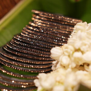 Set of six black glass bangles densely encrusted with champagne and rose gold crystal glitter, fanned out on a green banana leaf beside fresh white jasmine flowers in a wicker basket.