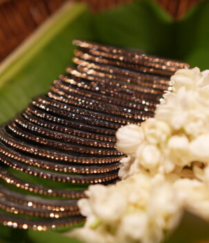 Set of six black glass bangles densely encrusted with champagne and rose gold crystal glitter, fanned out on a green banana leaf beside fresh white jasmine flowers in a wicker basket.