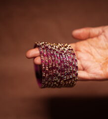 A stack of magenta and deep pink glass bangles densely covered in gold and silver crystal glitter, held in a woman's palm against a warm brown background.