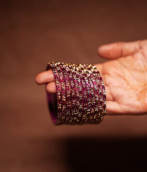 A stack of magenta and deep pink glass bangles densely covered in gold and silver crystal glitter, held in a woman's palm against a warm brown background.