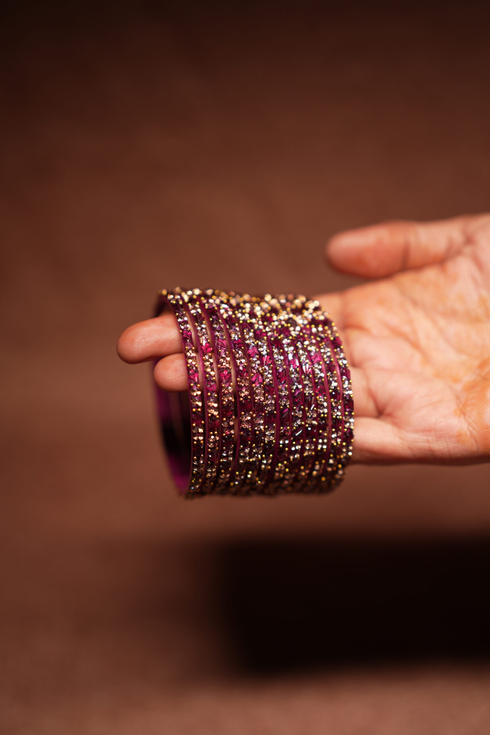A stack of magenta and deep pink glass bangles densely covered in gold and silver crystal glitter, held in a woman's palm against a warm brown background.