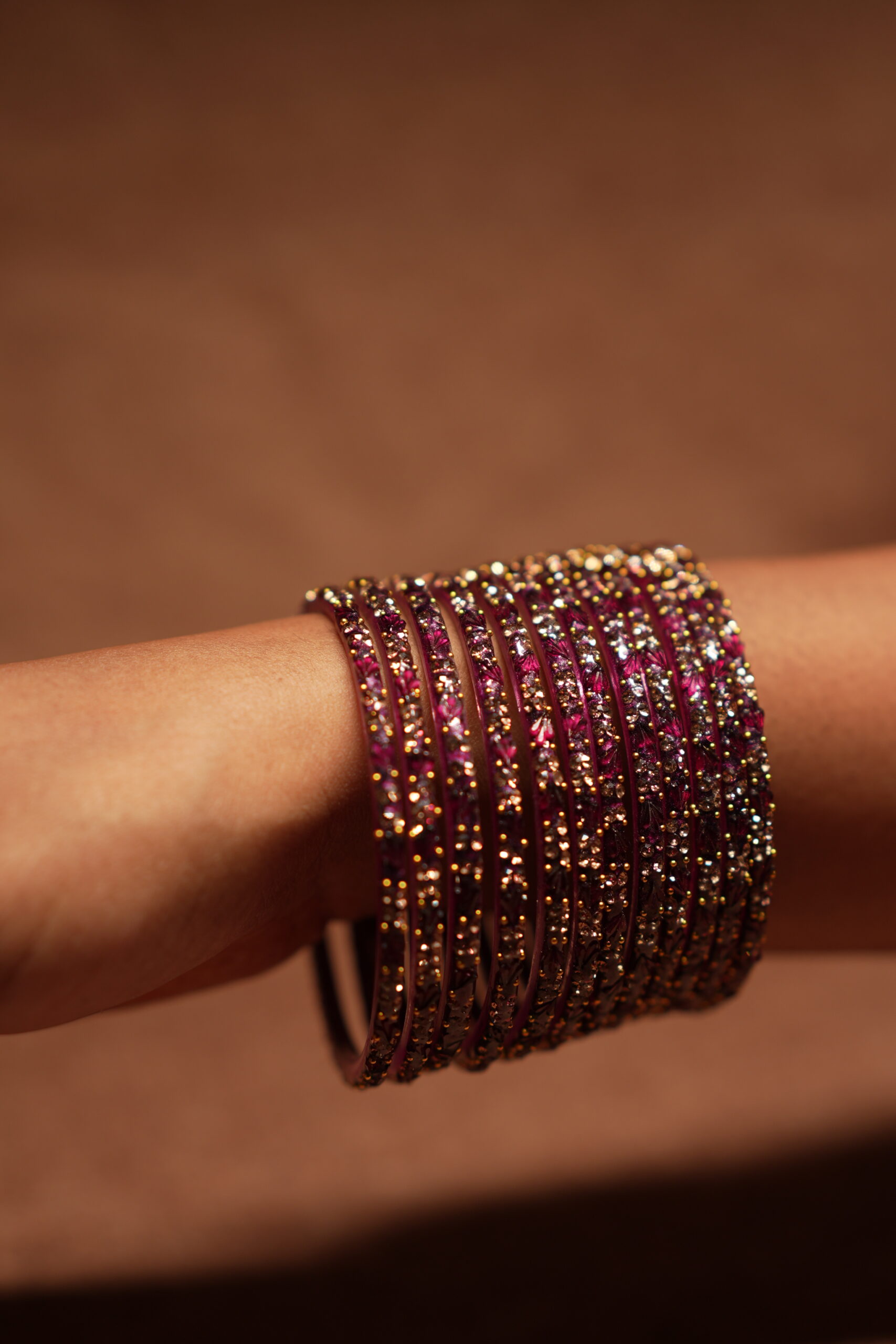 A stack of magenta and deep pink glass bangles densely covered in gold and silver crystal glitter, held in a woman's palm against a warm brown background.