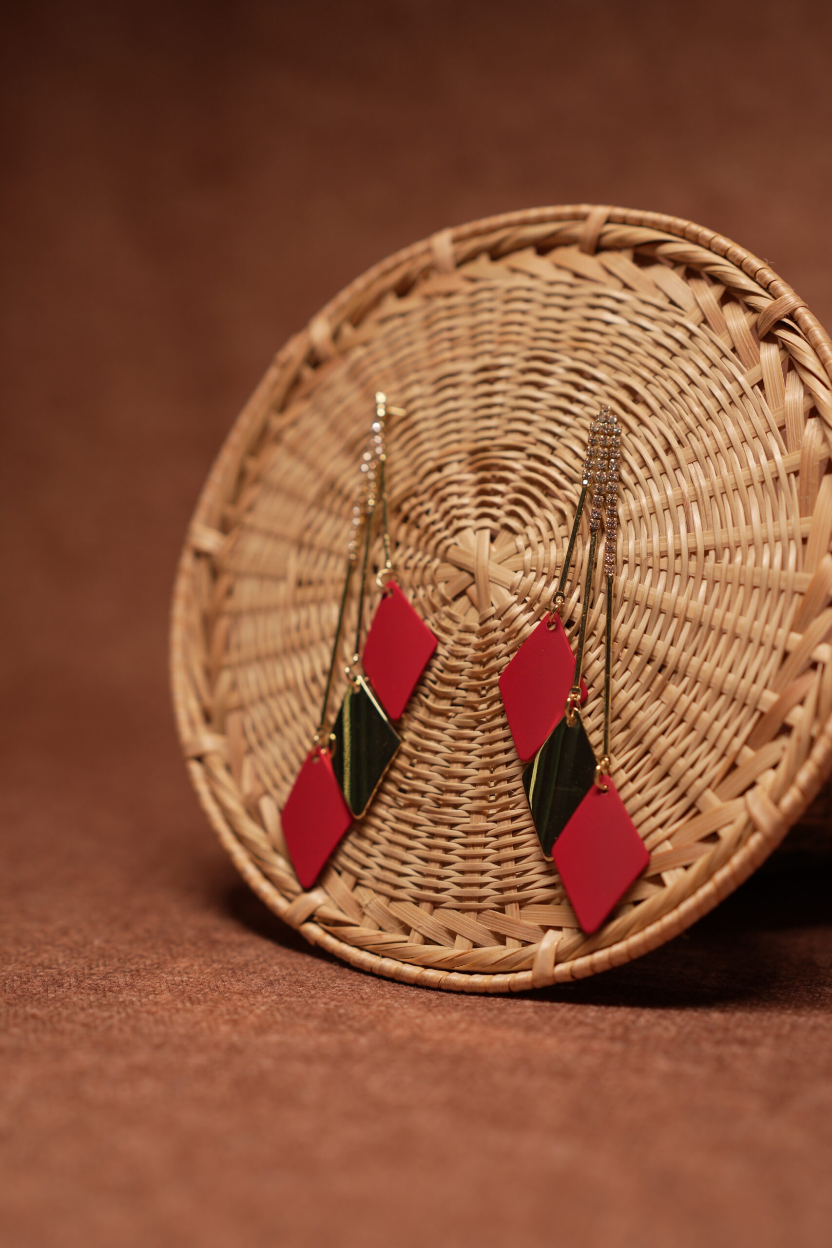 Red and green diamond shape layered drop earrings with long gold crystal chain tassel, displayed on a round wicker basket with warm brown background